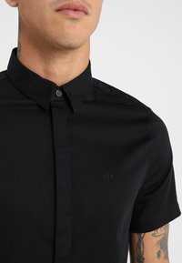 Man som bär en svart kortärmad skjorta med knappar och krage, synlig A|X-logga på br östet och tatueringar på höger arm.