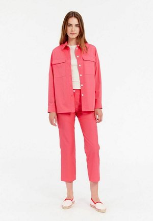 Chemise rose à manches courtes avec deux poches poitrine, assortie à un pantalon court et des espadrilles à semelle rouge. Texture lisse, coupe décontractée.
