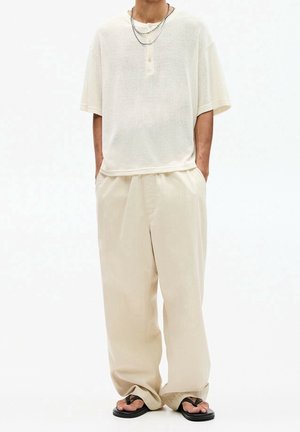 Person wearing loose cream-colored short-sleeve shirt and wide pants with black flip-flops, standing with hands in pockets.