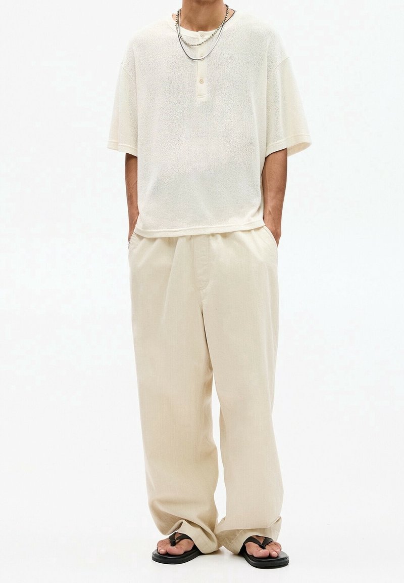 Personne portant une chemise ample à manches courtes de couleur crème et un pantalon large avec des tongs noires, debout les mains dans les poches.