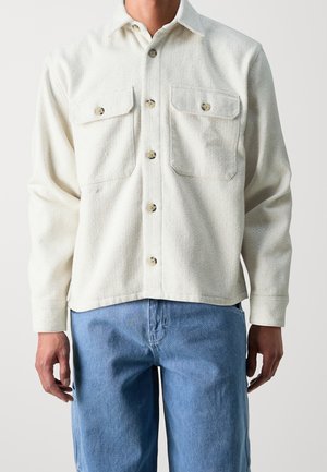 Mand iført en hvid struktureret skjorte med knapper og to forlommer samt blå jeans, stående foran en ensfarvet baggrund.