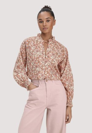 Frau mit Hochsteckfrisur trägt eine langärmelige Blumenbluse mit Volantkragen und hellrosa, hoch taillierte Hose, steht mit einer Hand in der Tasche.