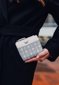 Kompaktní peněženka ve světle růžové a šedé barvě s květinovými vzory, s nápadným zipem a kovovým logo akcentem, držena v kapse černého kabátu.