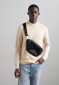 Schwarze Leder-Umhängetasche mit schlankem Design, verstellbarem Tragegurt und silbernen Beschlägen, getragen über einem hellbeigen Pullover und blauen Jeans.