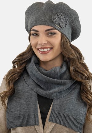 Berretto grigio a maglia con un accento floreale e sciarpa grigia con estremità a costine, entrambe realizzate in tessuto morbido, indossate da un modello con un cappotto beige.