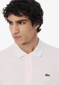 Polo shirt a righe rosa chiaro con colletto bianco e due bottoni, adornato da un piccolo logo di coccodrillo verde sul lato sinistro del petto.
