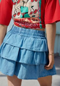 Denim lagdelt nederdel med tre lag; lys blå farve, elastisk talje og en slidt struktur. Matchet med en rød grafisk t-shirt ovenfor.
