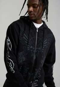 Junger Mann mit geflochtenem Haar trägt einen schwarzen Reißverschluss-Hoodie mit geflügeltem Totenkopf- und Rosendesign sowie dem Text "God Hates" auf dem Ärmel.