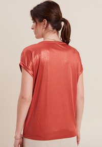 Korte mouwen, koraalkleurig topje van glanzende stof, met opgerolde manchetten en een ontspannen pasvorm. Het heeft een ronde halslijn en een subtiele textuur.
