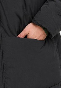 Melnā pūkainā jaka ar quilted tekstūru, kurai ir sānu kabata. Roka ieiet kabatā, parādot zaļu adītu aproci zem tās.