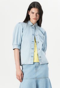 Young woman in a light blue denim jacket and matching skirt, with a yellow top underneath, gazes confidently at the camera.