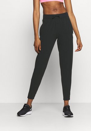 Femme portant un pantalon de jogging noir, des baskets noires et un soutien-gorge de sport rose, debout, jambes légèrement écartées, sur un fond neutre.