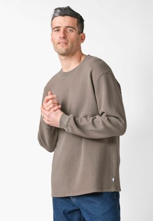 Uomo con capelli corti e scuri che indossa una maglia a maniche lunghe color taupe e jeans blu, in piedi con le mani giunte davanti al petto su sfondo bianco.