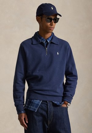 Muž nosiaci námornícky modrý sveter so štvrťzipsom cez modrú károvanú košeľu, tmavé džínsy, námornícku čiapku, slnečné okuliare a náramkové hodinky stojiaci na neutrálnej pozadí.