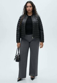 Geacă puffer neagră cu quilt-ing orizontal, purtată cu pantaloni largi gri cu dungi fine. Accesoriile includ o geantă mică neagră și pantofi cu vârf ascuțit.