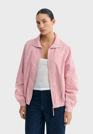 Frau mit dunklen Haaren trägt eine rot-weiß vertikal gestreifte Jacke mit Reißverschluss über einem weißen Tanktop und dunkelblauen Jeans.