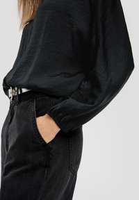 Schwarze Bluse mit langen Ärmeln aus einem weichen, strukturierten Stoff und lässiger Passform, kombiniert mit hoch taillierten schwarzen Jeans mit seitlichen Taschen.
