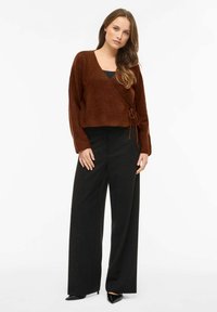 Sweter zapinany w kolorze rdzy z detalem wiązania, wykonany z teksturowanej tkaniny, zestawiony z czarnymi spodnimi o szerokich nogawkach oraz czarnymi, spiczastymi butami.