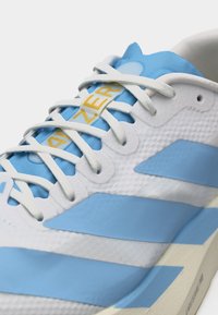 Gros plan sur une chaussure de sport blanche et bleu clair avec des lacets blancs et un texte jaune "AZERO" sur la languette bleue.