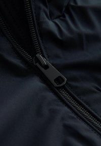 Sort nylon stof med et nærbillede af en robust lynlås. Lynlåsen har en mat sort trækhank og er syet ind i stoffet uden synlige sømme.