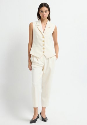Cream sleeveless vest with five gold buttons, tapered design, paired with matching cream trousers and black heeled shoes.