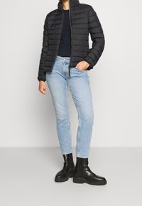 Černá péřová bunda s vysokým límcem, kombinovaná se světlomodrými džínami rovného střihu a černými robustními botami. Hladký povrch a zipové zapínání.