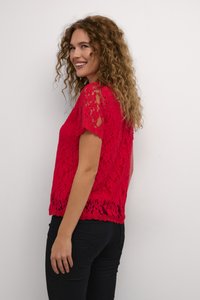 Haut en dentelle rouge à manches courtes, présentant des motifs floraux et un ourlet festonné. Assorti avec un pantalon noir cintré, sur un fond blanc.