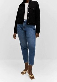 Giacca di jeans nera con due tasche sul petto, abbinata a jeans azzurri aderenti alla caviglia. Modello indossa sandali gialli con cinturini, in piedi su uno sfondo neutro.