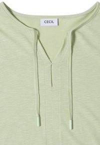 Chemise vert clair avec une ouverture en V et des cordons, avec une étiquette blanche portant le nom de la marque "CECIL" au niveau de l'encolure.