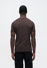 Καφέ T-shirt από βαμβάκι, με στρογγυλή λαιμόκοψη και κοντά μανίκια. Η πίσω όψη αποτυπώνει μια λεία υφή και ελάχιστο branding στη λαιμόκοψη.