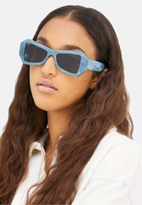 Occhiali da sole geometrici blu con montatura spessa e lenti scure, caratterizzati da una texture marmorizzata e design angolare.
