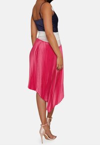 Plissierter Midirock in Pink mit einem asymmetrischen Saum und weißem Bund. Kombiniert mit einem figurbetonten Oberteil in Marineblau und beigen Riemchen-Heels.