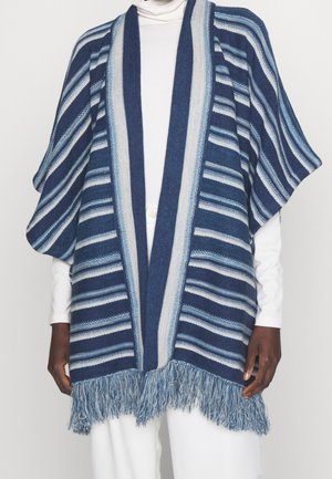 Personne portant un châle à rayures bleues et blanches ouvert devant avec un ourlet à franges, par-dessus un col roulé blanc et un pantalon.