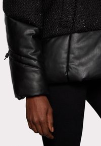 Černá vyp padded bunda s texturovanou horní částí, hladkou koženou spodní částí a elastickými manžetami. Vyznačuje se jemným leskem a přiléhavým střihem.