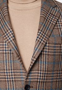 Brązowa marynarka w kratę z niebieskimi akcentami, z wełnianego materiału, z jednym czarnym guzikiem i beżowym golfem pod spodem. Widoczny szczegół tekstury.