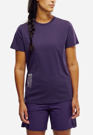 Femme aux cheveux tressés portant un t-shirt violet uni à manches courtes et un short violet assorti, debout devant un fond blanc.