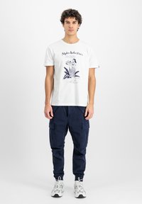 Tricou alb din bumbac cu un print grafic ce o înfățișează pe o femeie ținând fructe, combinat cu pantaloni cargo bleumarin, cu buzunare laterale și manșete elastice.