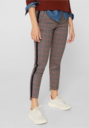 Vrouw draagt enkelhoge pied-de-poule broek met zwarte zijstrepen, witte sneakers, spijkershirt en bruine trui.