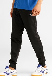 Pantalons de sport noirs avec un design fuselé, dotés de poches latérales et d'une taille élastique. Assortis à des chaussures de sport blanches et bleues.