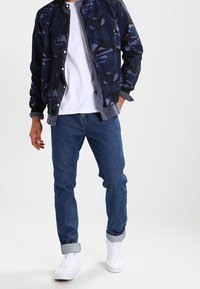 Persona con chaqueta bomber de camuflaje azul, camiseta blanca, jeans azules doblados y zapatillas blancas, de pie con una mano en el bolsillo.