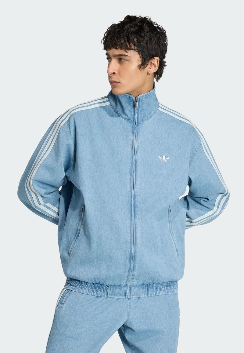Jonge man met donker krullend haar, gekleed in een lichtblauw Adidas jack met rits en witte strepen, en bijpassende broek, kijkt opzij.
