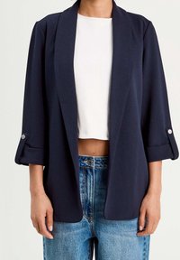 Blazer bleu marine en tissu doux, doté d'un col châle, de manches retroussées trois-quarts avec boutons en accent, et d'une coupe ouverte à l'avant.