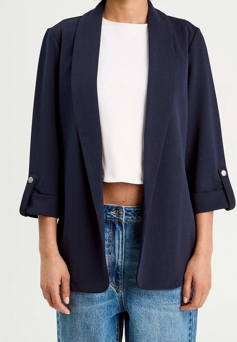 Blazer bleu marine en tissu doux, doté d'un col châle, de manches retroussées trois-quarts avec boutons en accent, et d'une coupe ouverte à l'avant.