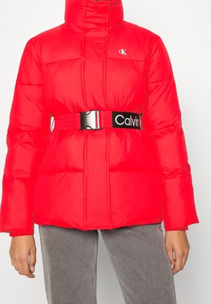 Rød dunjakke med høj krave, sort justerbart bælte med hvid "Calvin" tekst, båret med grå bukser, hænder afslappet ved siderne.