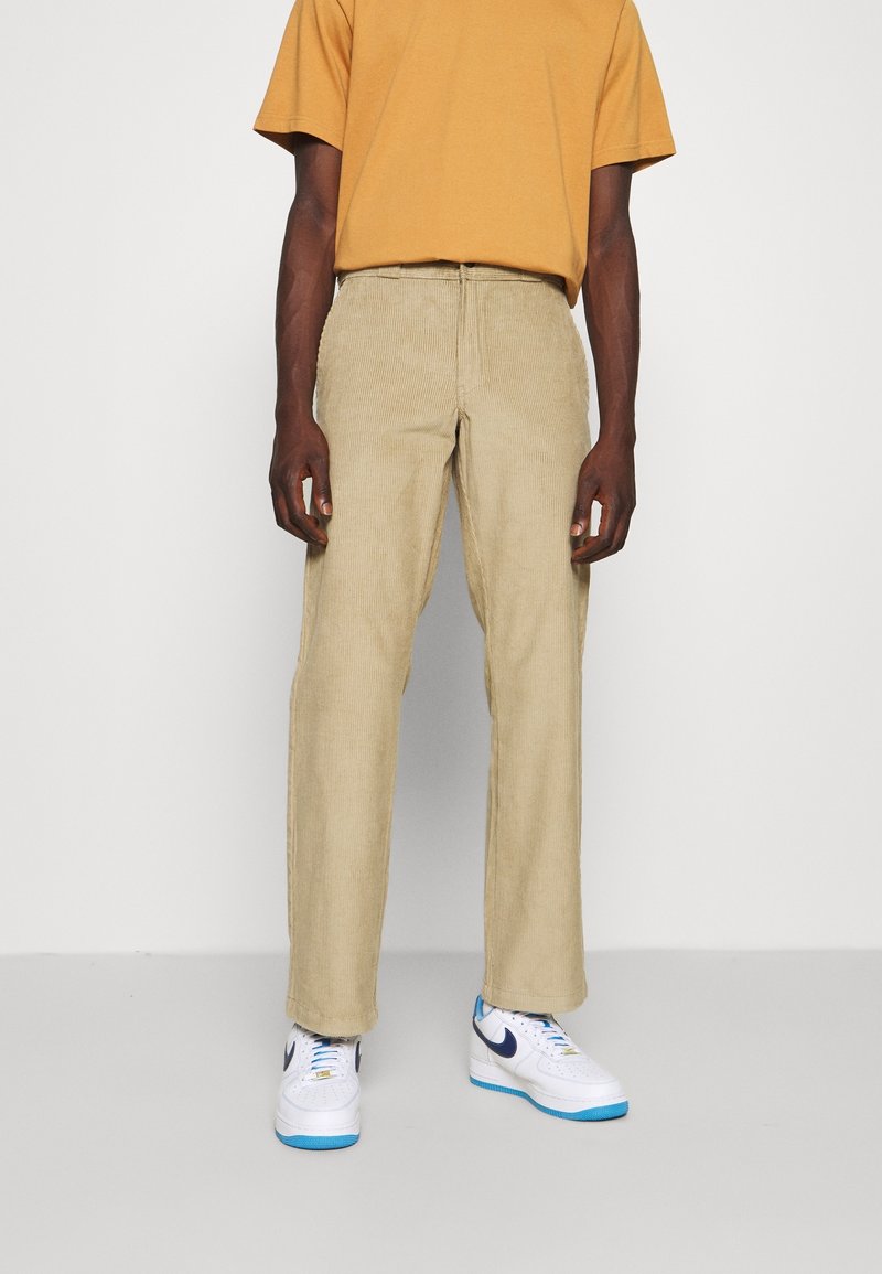 Beige kordflöjtsbyxor med avslappnad passform, med en texturerad yta. I kombination med en senapsgul t-shirt och vita sneakers.