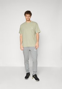 Olijfgroene katoenen T-shirt met een borstzak, gecombineerd met grijze trainingsbroek met een logo. Zwarte sportschoenen met groene accenten.