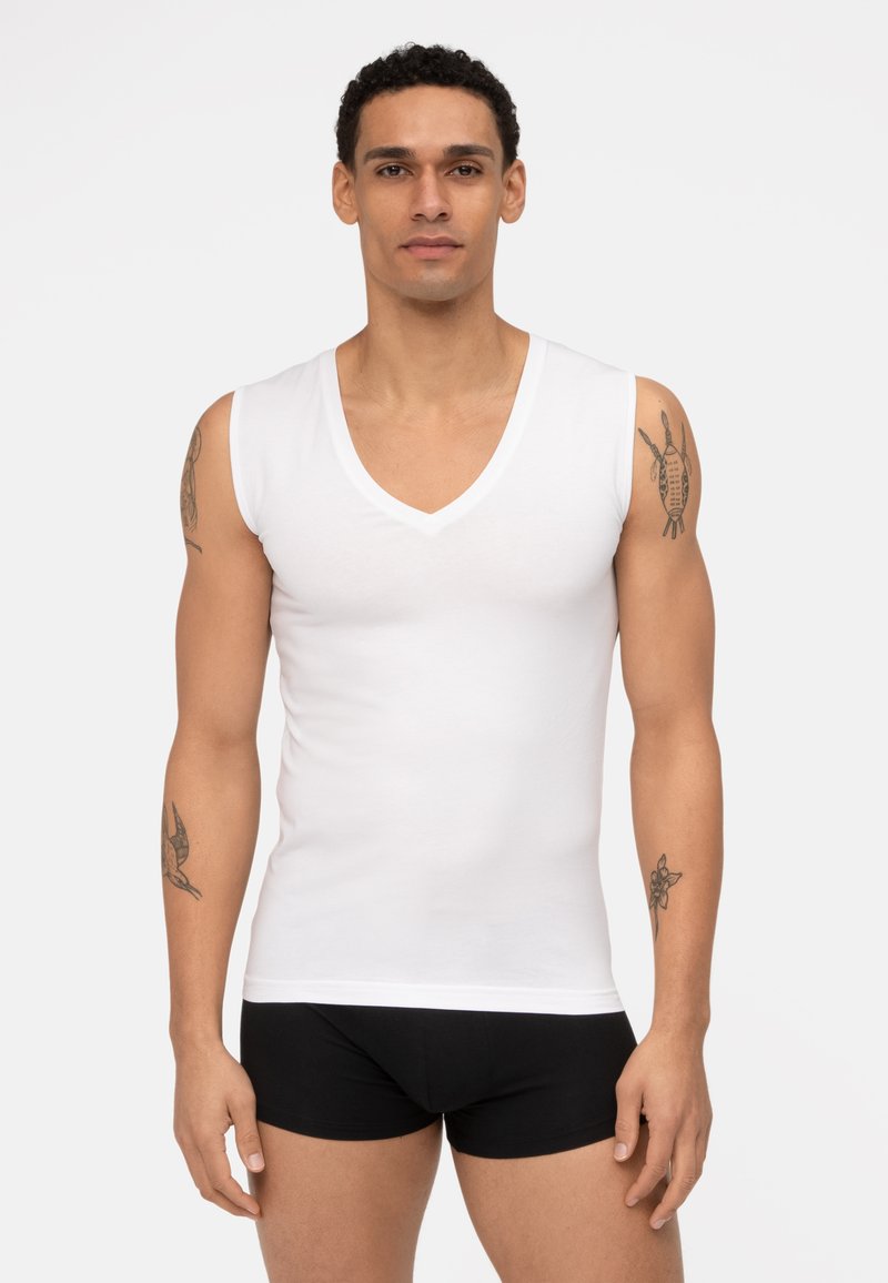 Witte katoenen mouwloze V-hals t-shirt, met een gladde textuur, fitted ontwerp, gedragen met zwarte shorts, met zichtbaar armtattoo's.