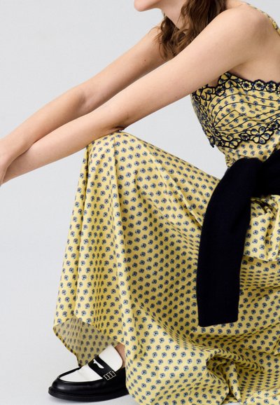 Maxi dress giallo con motivo a fiori e rifiniture in pizzo nero. Scarpe slip-on bianco e nero completano il look.