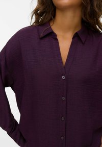 Chemise violette à boutons avec un tissu doux et texturé, dotée d'un col et de six boutons assortis sur le devant.