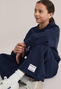 Tumši zils džemperis un bikses komplekts, izgatavots no mīkstas auduma. Biksēs ir logotipa uzlika un ribas aproces. Balti potīšu zeķes un sporta apavi.